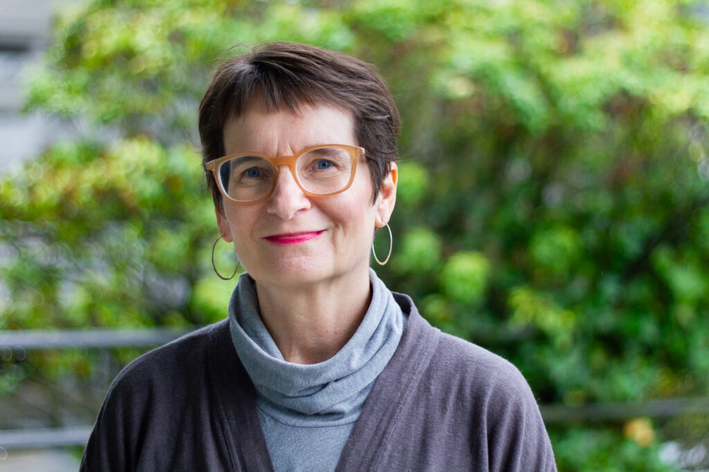 Lois Klassen, a white woman with short brown hair and glasses is smiling for the camera with blurred trees in leafe in the background. She is wearing hoop earrings and grey turtleneck top.