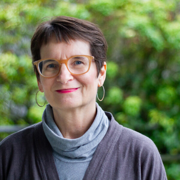 Lois Klassen, a white woman with short brown hair and glasses is smiling for the camera with blurred trees in leafe in the background. She is wearing hoop earrings and grey turtleneck top.