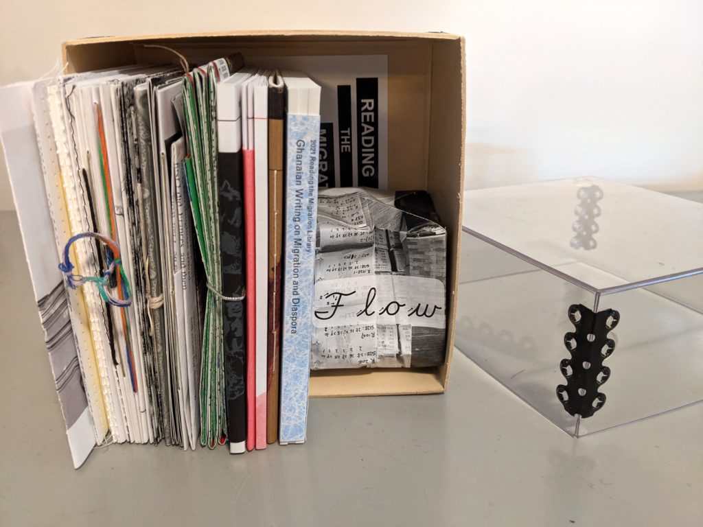 An archive box with a tan coloured base and clear plexi-glas top is seen on its side with the contents standing as they would on a book shelf. A plexiglass lid is lying next to it on a grey surface. It contains the full set of RML books which are various colours, thicknesses and textures. One is folded into the shape of an orgami balloon.