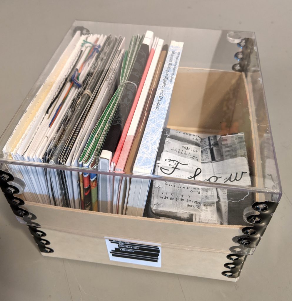 An archive box with a tan coloured base and clear plexi-glas top is seen from above. It contains the full set of RML books which are various colours, thicknesses and textures. One is folded into the shape of an orgami balloon.