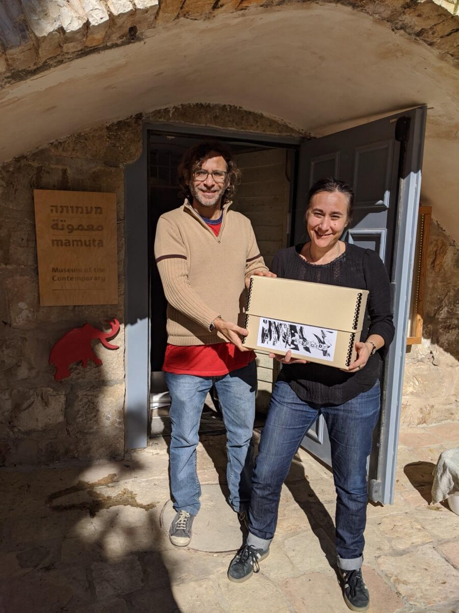 Two people are holding an archive box in the sunlight on the stoop of Mamuta Art and Research Center, Jerusalem.