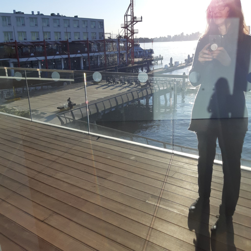 A woman is shown on right taking a photo of her own reflection in a window. Another reflection of herslef appears in her shadowy image. In the background there is a wooden peir and a dock. Burrard Inlet is seen in the background.