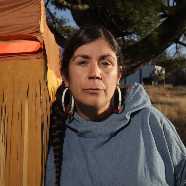 Tania Willard is pictured outdoors with a tree and buildings in the background. Willard is looking directly at the camera and leaning on a structure with a cover of leather strips that cover Willard's hand. Willard is wearing beaded hoop earings and has a long dark braid over their right shoulder.