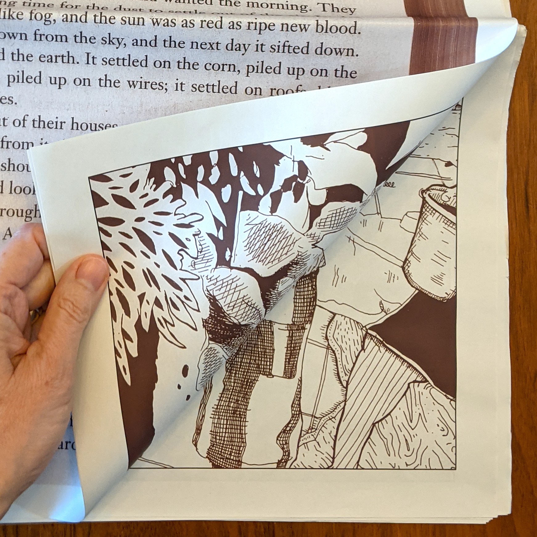 A hand is seen turning the page on Juan Cisnero's newspaper titled Grapes of Steinbeck. The pages are off-white and the ink is brown. The page being turned has printed text on the facing side but comic images are revealed as the page is turned to the paper's inside surfaces.