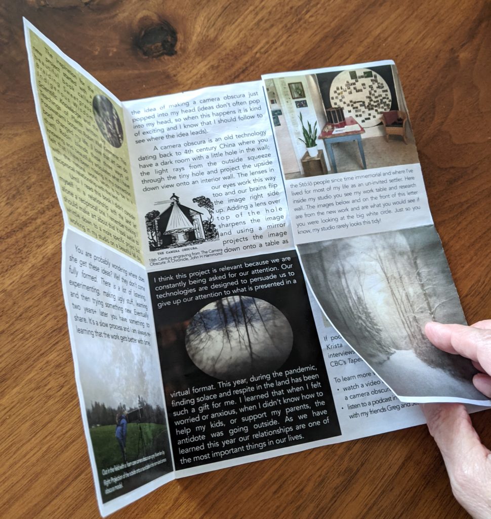 A full colour flyer is shown lying on a wooden table. Part of a hand is seen unfolding the flyer to reveal that all sides are printed with text and camera obscura photos.