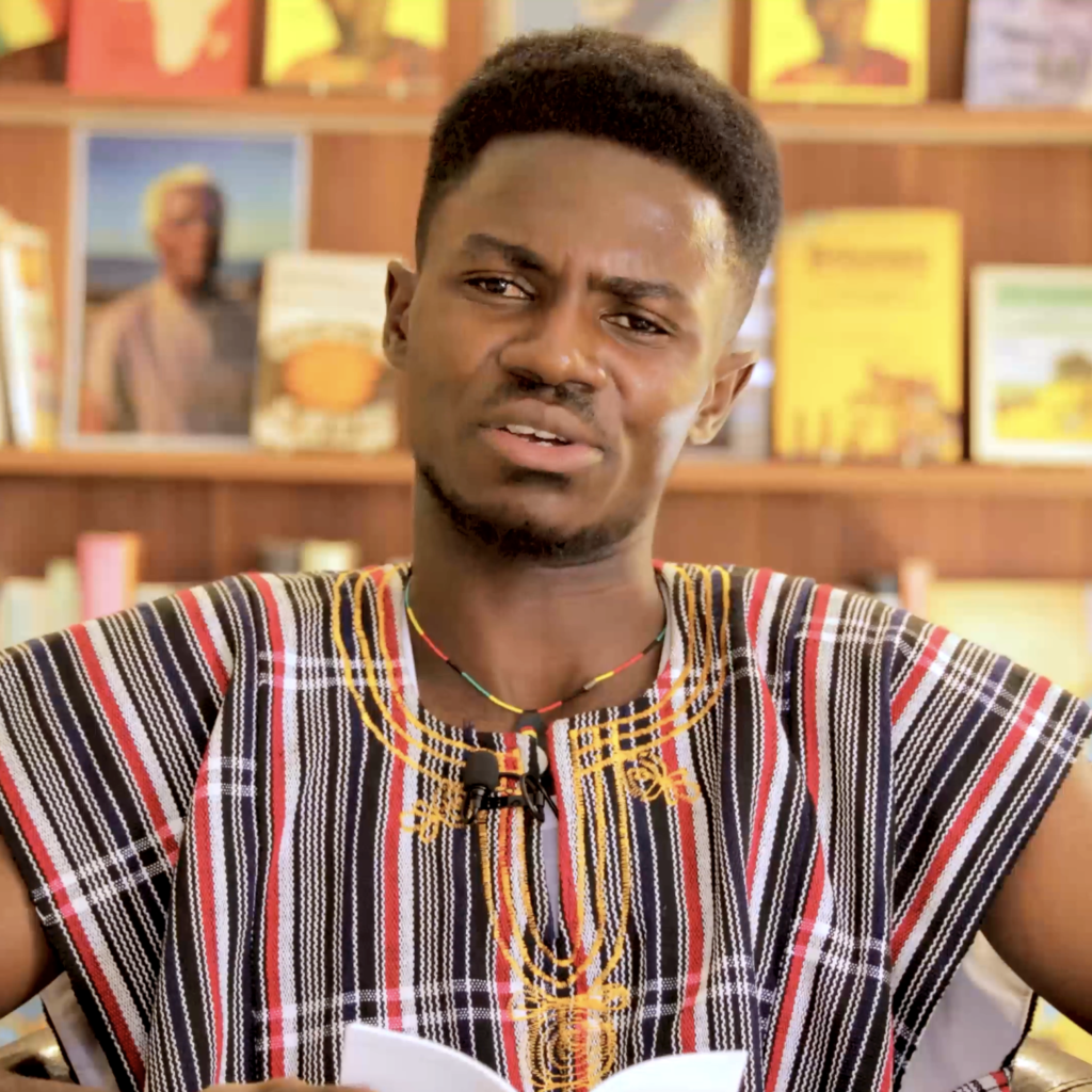 A man with black hair, brown skin, and Ghanaian woven and embroidered shirt is seen facing th camera, reading from a small book. There are library shelves of books behind him.