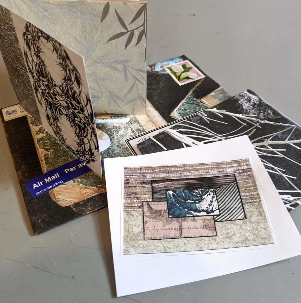 Collaged card, envelope and small book are on a grey surface. These items are each decorative combinations of textured and coloured papers in grey, blue and black tones.