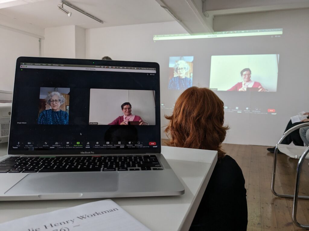 Lois Klassen's laptop displays Ginger Mason (L) and Lois Klassen (R) in conversation using zoom. In the background, the screen is projected on the wall. In between there is the back of an audience member's head. They have dark red hair.