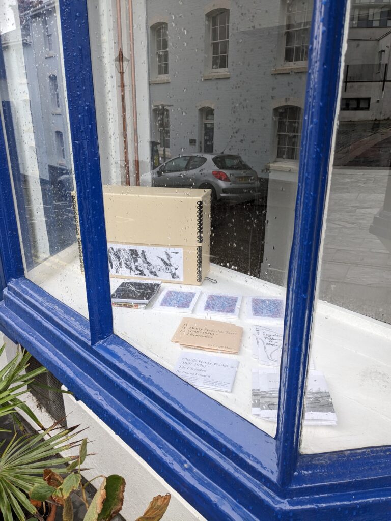 Looking through the rain-marked bay window of 37 Looe Street, RL Box#14 and various books from the RML project are seen arranged on the interior window sill. The exterior window frame is painted in bright blue. 