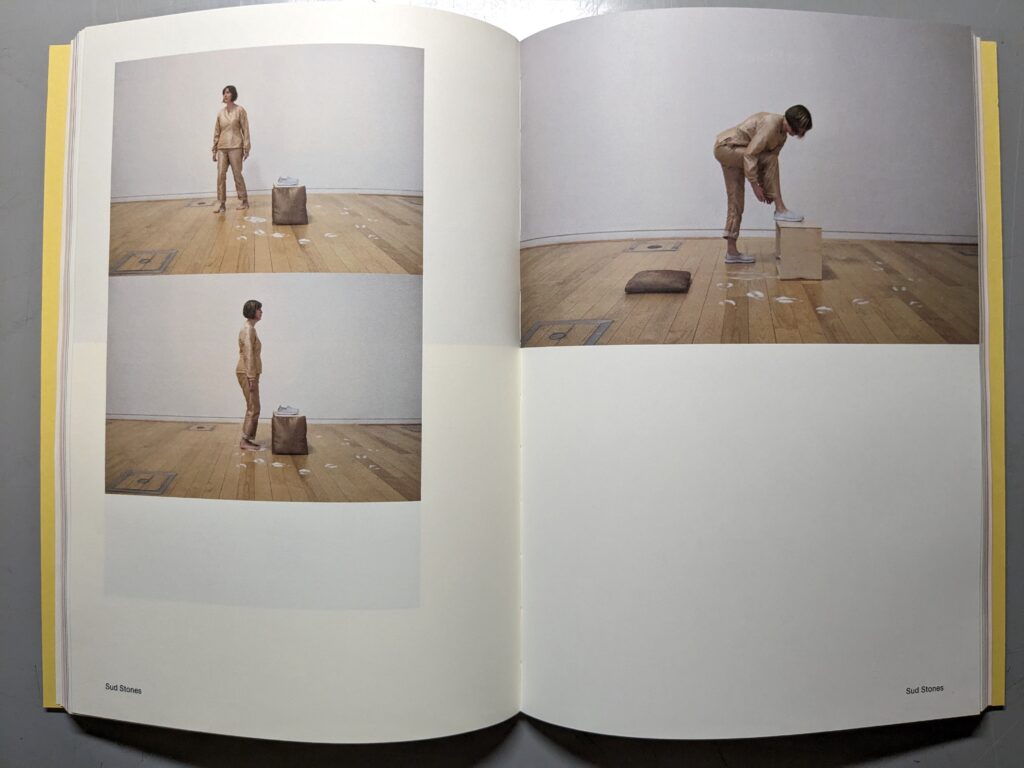 An open spread from the book "Four Hundred and Twenty-nine Significant Moments: Documenting an Artist's Research and Processes" by Lisa Watts. The pages include 3 images of the artist in solo performances: a single figure wearing a shiny gold-coloured pantsuit and with brown hair in a bob cut, in a white walled room with polished wood plank floor. The artist walks and stands near a box with a brown cushion and white shoes. In one photo she is putting the shoes on. There are white powdered foot prints on the floor.