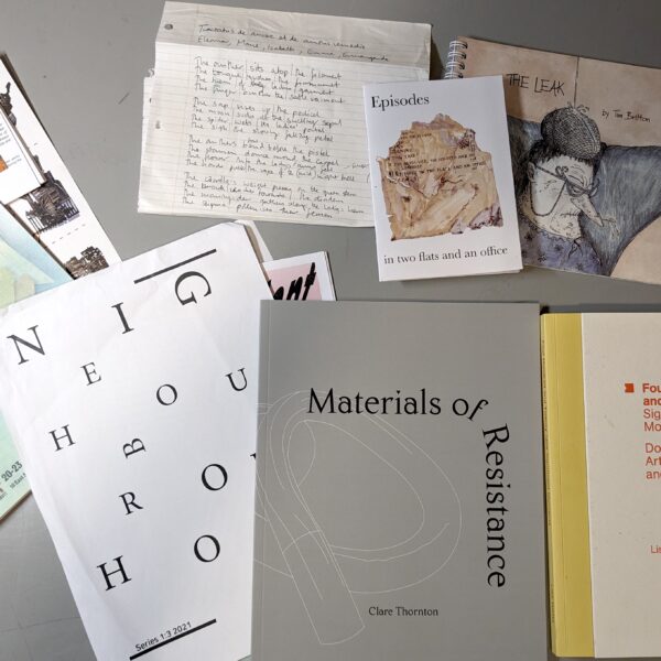 A birds eye view of the artist books and ephemera that were received in exchange for books in the Reading the Migration Library exchange event at 37 Looe Stree in Plymouth, UK on September 19, 2023. In clockwise order starting top left, colour printed cards and poster from "Ruptured Domesticity" project, a handwritten lyric sheet on ruled paper by Mo Bottomley, a small colou-printed 'zine titled "Episode in two flats and an office" by Sef Penrose, a colour printed and coil-bound picture book called "The Leak" by Tim Britton, a thick, hardcover artist monogram titled "Four Hundred and Twenty-nine Significant Moments: Documenting an Artist's Research and Processes" by Lisa Watts, a grey artist's catalogue called "Materials of Resistance" by Clare Thornton, and a sheaf of loose printed papers making up a publications called "Neighbourhood 1. (Series 1:3 2021) by Merrydith Russell.