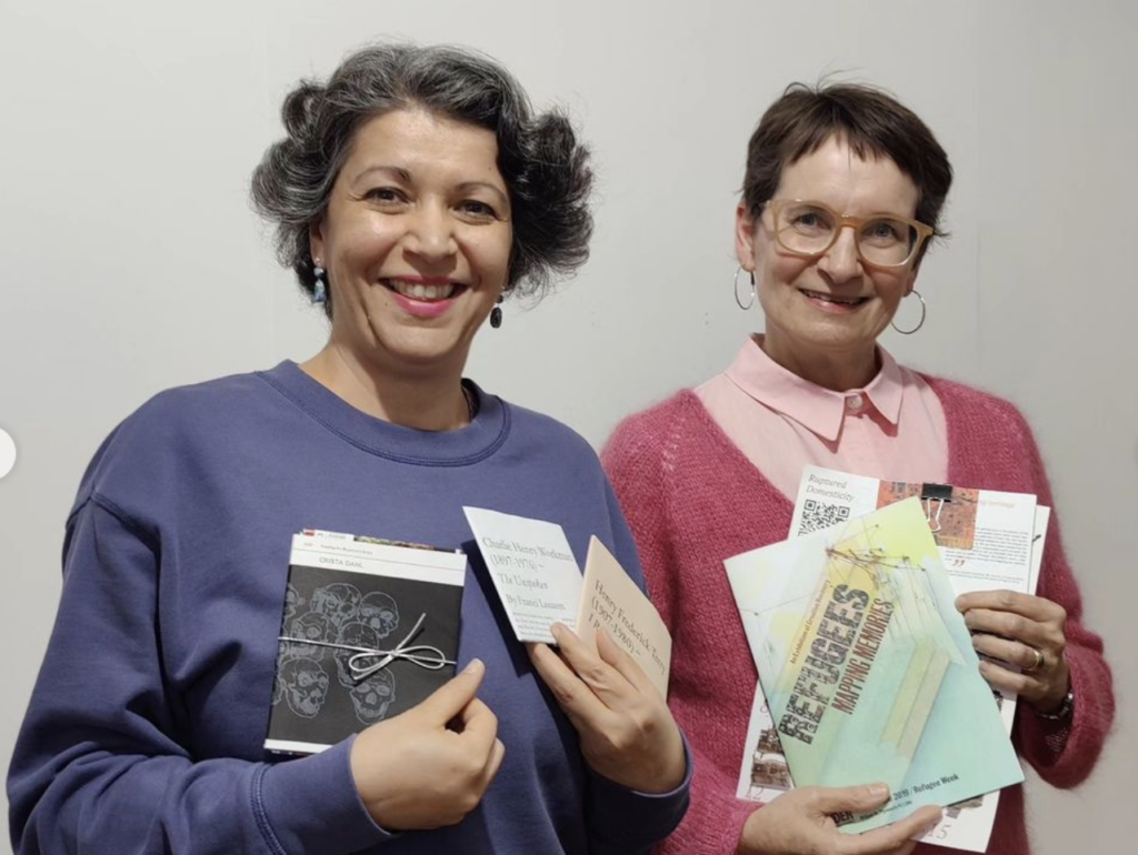 Sana Murrani (L) is holding 3 RML publications. Sana is wearing a purple collarless pullover. Sana has loosely curled salt and pepper coloured short hair. Lois is wearing a pale pink button-down shirt with a darker pink v-necked sweater. Lois has short dark brown hair and glasses.