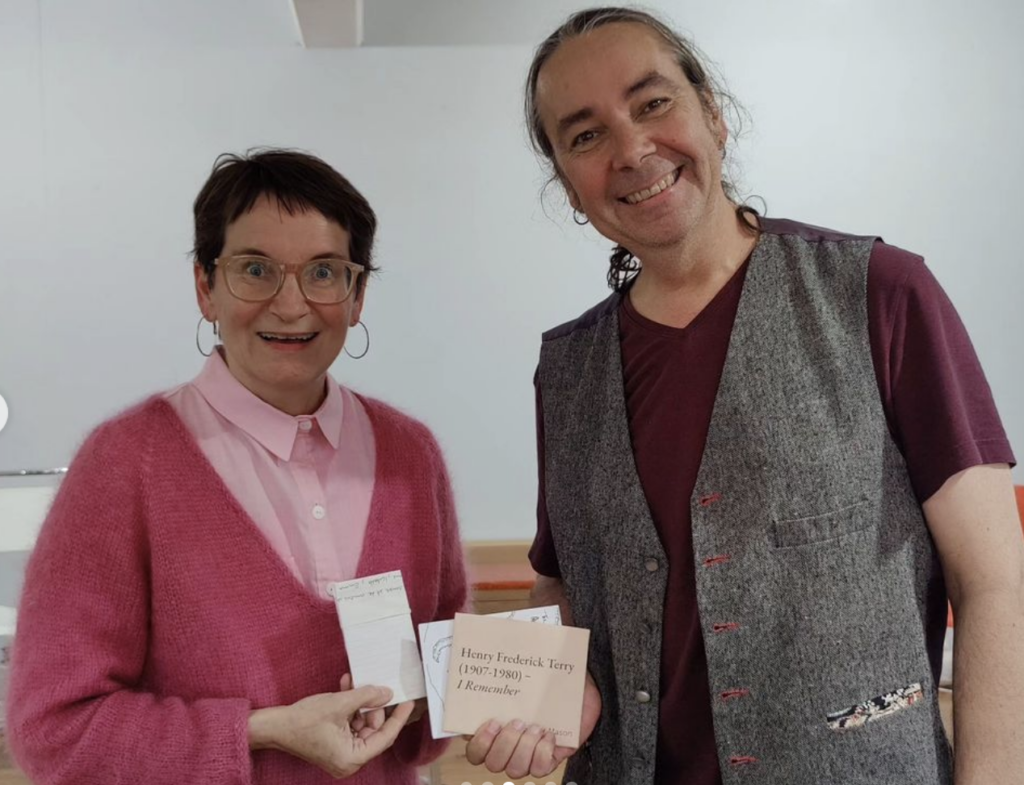 Lois Klassen (L) holds a folded paper with song lyrics and Mo Bottemley (R) holds copies of RML books. Lois is wearing a pale pink button-down shirt with a darker pink v-necked sweater. Both have light-coloured skin. Lois has short dark brown hair and glasses. Mo has hair pulled back in a pony tail. He is wearing a grey vest over a wine-coloured collarless shirt. Lois and Mo are smiling excitedly.