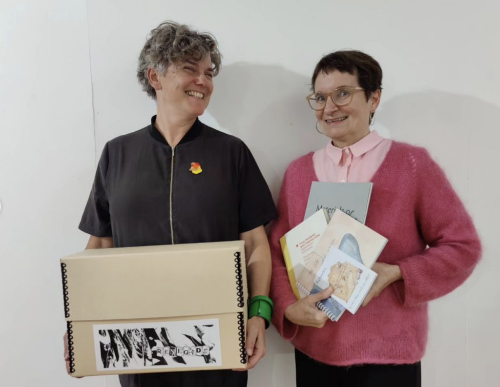 Angela Piccini (L) holds RL Box#14 While Lois Klassen (R) holds a handful of artist books that have been traded for the box. Angela is wearing dark, shirt-sleaved coveralls with a front zipper. She has salt and pepper colored hair. Lois is wearing a pale pink button-down shirt with a darker pink v-necked sweater. Both have light-coloured skin. Lois has short dark brown hair and glasses.