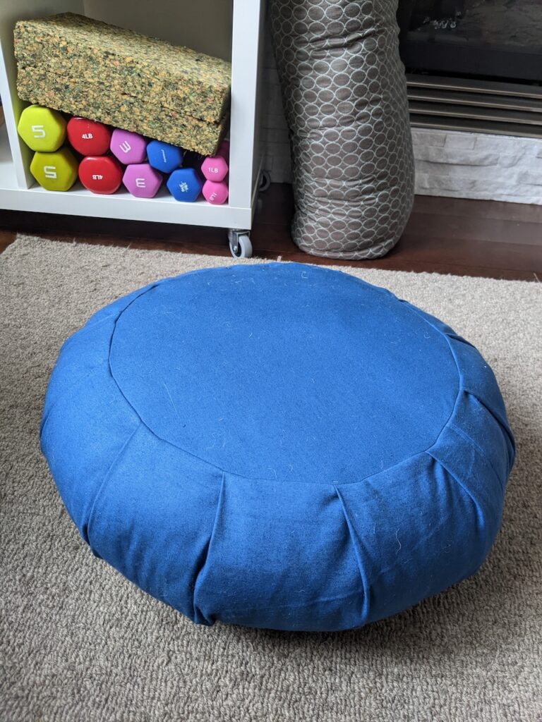 A round meditation cushion is on a carpetted floor. In the background, exercise weights (dumbbells and other yoga cushions and blocks) are seen in the background. 