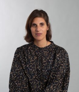 Sarah Shamash is smiling and facing the camera in a conventional studio portrait. There is a featureless grey background. She has shoulder-length brown hair, dark eyes, and light brown skin. She is wearing a flowered blouse.