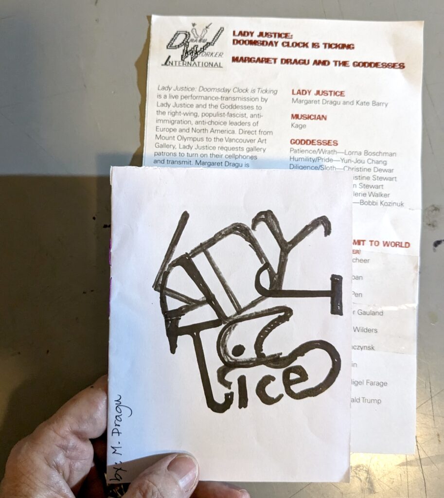 A hand is holding a small 'zine by M. Dragu. The abstracted letters on the cover spell in black pen, "Lady Justice". Behind the 'zine a brochure describes activities from the Lady Justice performances by Margaret Dragu.