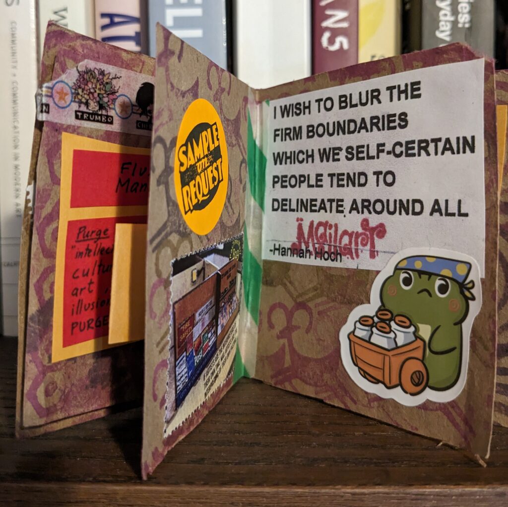 A tiny book is standing open on a shelf. It is made of brown paper and filled with stickers, quotes, and rubber stamped images.