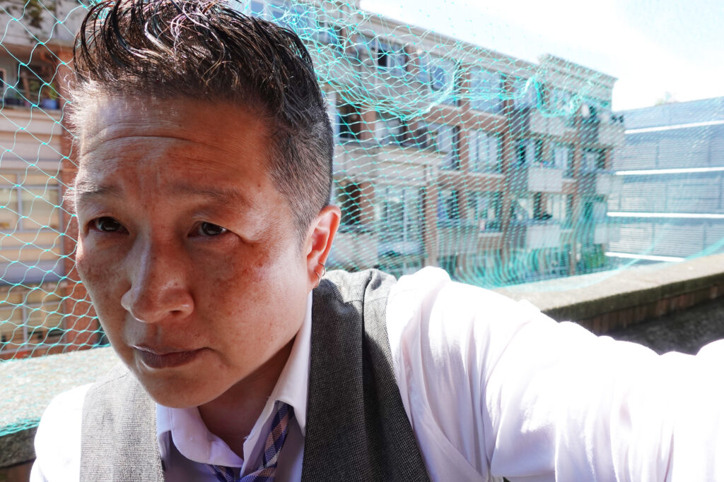 Candie Tanaka is pictured outdoors with sun shining on their shoulder and apartments in view behind a green plastic mesh. They are wearing a white shirt, grey vest, and neck tie. They have short cropped hair and have light brown skin tones.