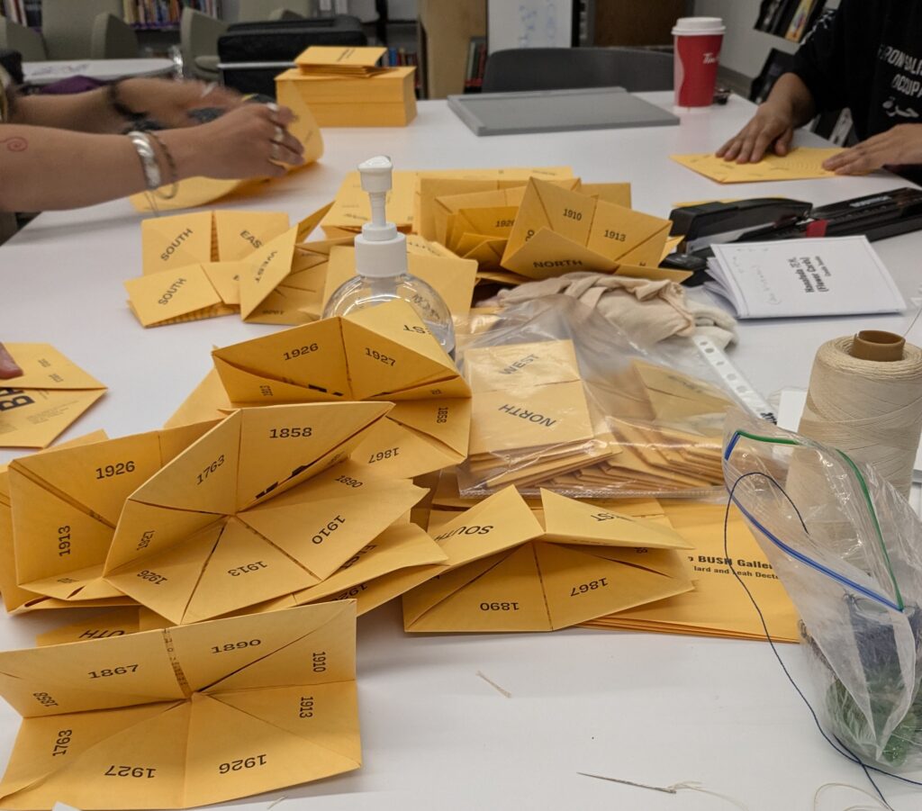 An view of the folded "fortune tellers" that are part of Directions to BUSH Gallery by Tania Willard and Leah Decter. They are in a pile and there are people's hands seen in the act of folding them. The fortune tellers are printed in dark purple ink on goldenrod yellow paper. 