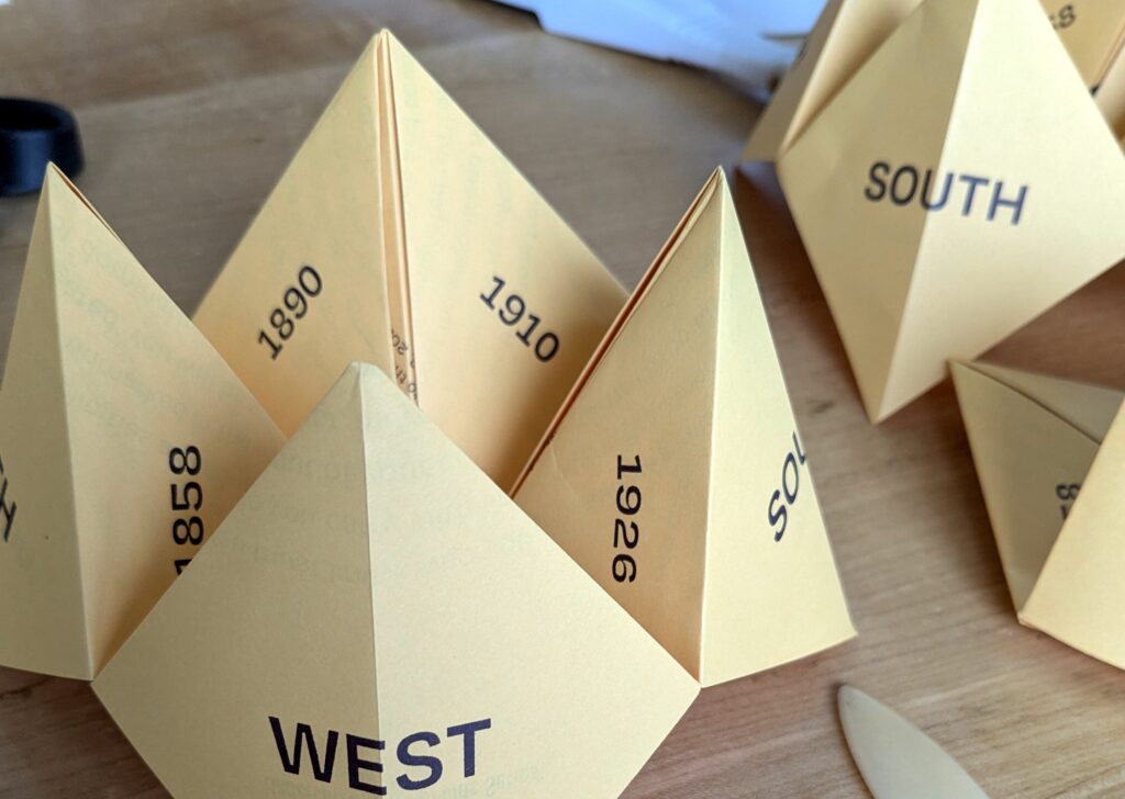 Two "fortune tellers" that are part of Directions to BUSH Gallery by Tania Willard and Leah Decter are seen folded and sitting on a table. The fortune teller is printed in dark purple ink on goldenrod yellow paper. 