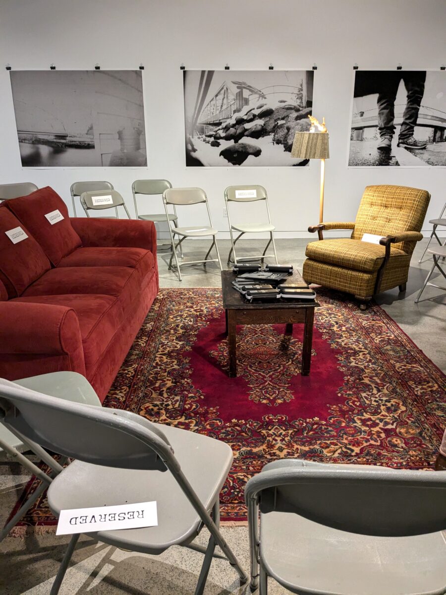 The exhibition space for SAFE is empty of people but includes a couch, arm chairs and lamps surrounding a coffee table with zines and Naloxone kits on it. The furniture is on a patterned rug. There are folding chairs surrounding the living room furniture, waiting empty for people to arrive for the exhibition opening. Many of the chairs and all of the upholstered furniture have the sign "reserved" on them.