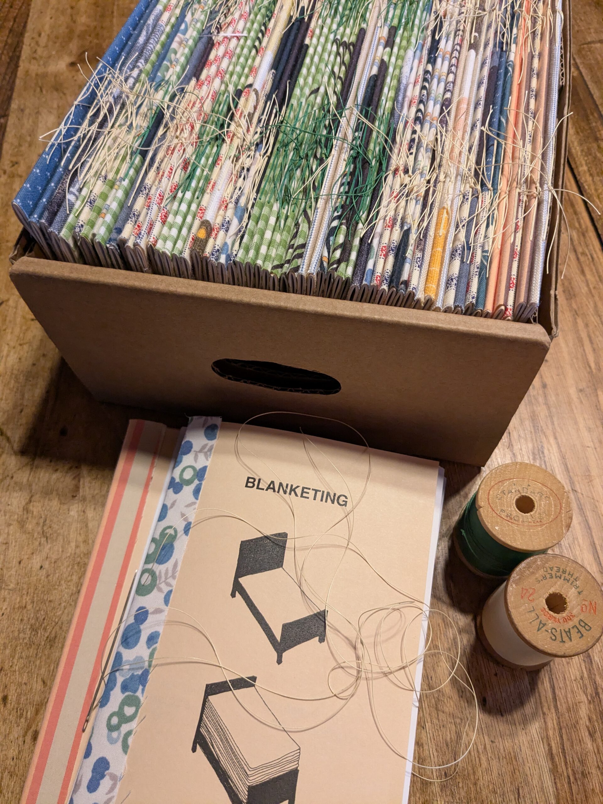 A few copies of the publication "Blanketing" is on a wooden table with two wooden spools of thread. Behind is a cardboard box stuffed with more copies. They have strips of colourful fabric along the spine and the thread binding is exposed.